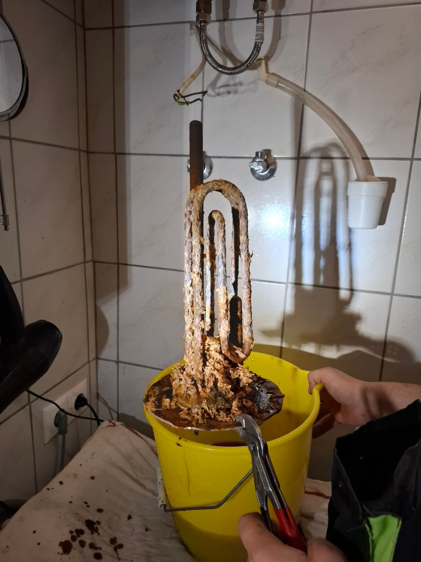 Erősen elvízkövesedett bojler fűtőbetét kiemelve a tartályból, vödör felett
